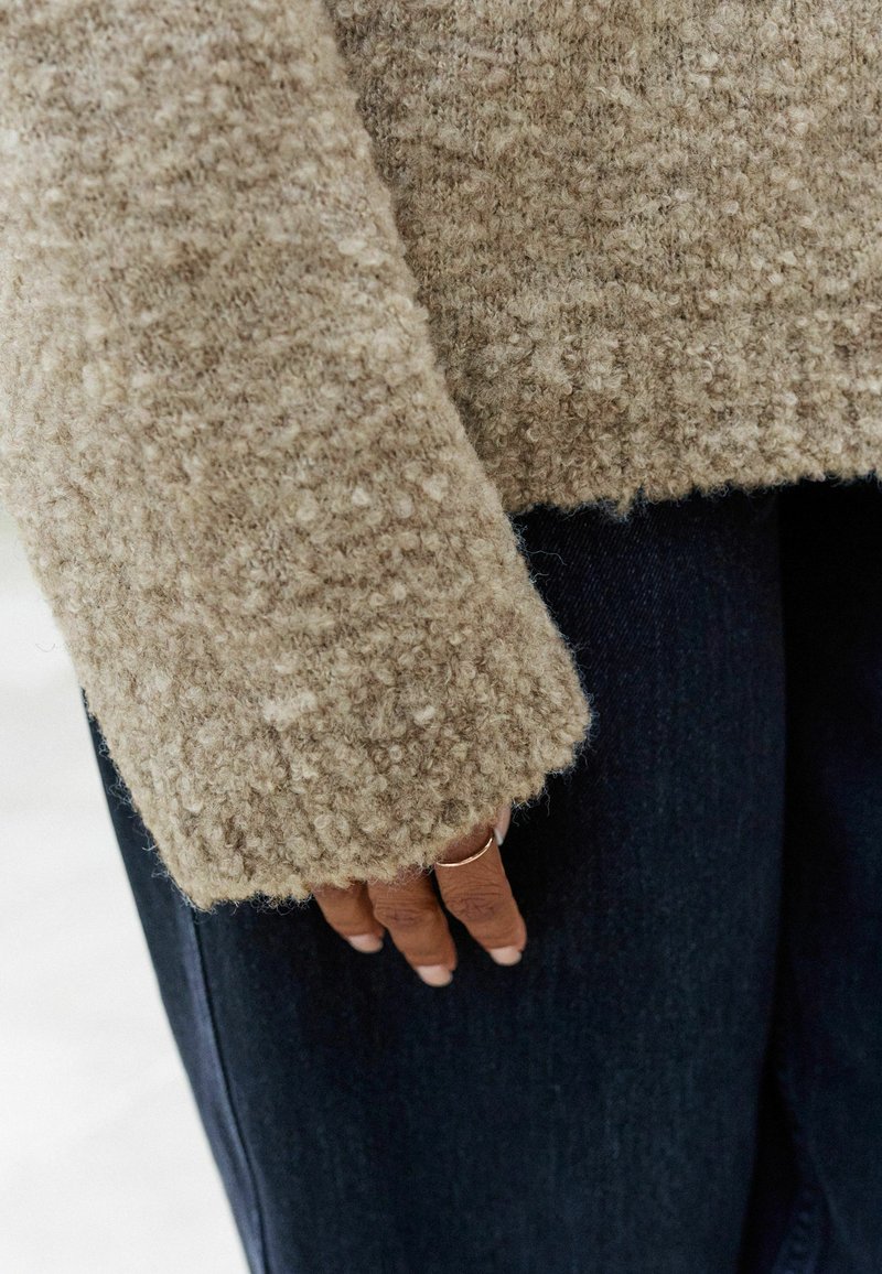 Beige texturerad tröja med lös passform, som har mjukt, fluffigt tyg och rå fåll, bärs över mörk denimjeans, hand med ring synlig.