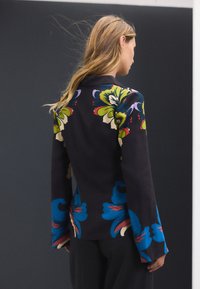 Blazer noir à motif floral avec des motifs colorés en vert, bleu et rouge, coupe ajustée et manches longues, visible de dos.