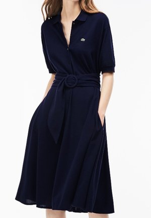 Vrouw die een marineblauwe jurk met kraag draagt, korte mouwen, knoopsluiting aan de voorkant, zijzakken en een bijpassende stoffen riem met een ronde gesp.