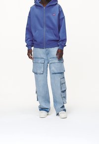 Felpa blu con zip, dotata di tasche frontali e un piccolo logo, abbinata a pantaloni cargo azzurri chiari con multiple tasche laterali e una vestibilità ampia.