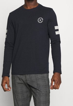 Homme portant un t-shirt à manches longues noir avec un logo circulaire blanc "Pier One" sur la poitrine et deux bandes blanches sur chaque manche, assorti à un pantalon à carreaux gris.