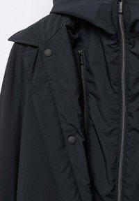 Abrigo negro impermeable con capucha, cierre de cremallera y botones de presión, con una textura suave y un diseño elegante.