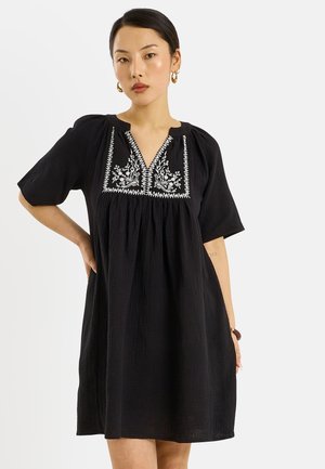 Femme portant une robe noire ample à manches courtes avec une broderie florale blanche sur la poitrine, posant avec une main sur la hanche.