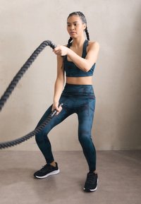 Top corto sportivo a fantasia teal e leggings a vita alta, abbinati a sneakers nere. La modella si allena con una corda da battaglia nera.