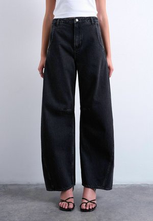 Person iført løstsiddende sort denimjeans med høj talje, kombineret med sorte sandaler med åbne tæer og stropper, stående på en grå gulv.