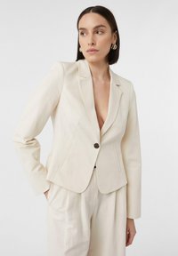 Jasno beżowa marynarka dopasowana z klapami, zapinana na jeden guzik, z dwoma przednimi kieszeniami. Gładka tkanina z wyraźnie zarysowanymi ramionami.