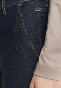 Nahaufnahme von dunkelblauen Denim-Jeans mit orangefarbenem Stich und einem beigen Langarmhemd, dessen Ärmelbund das GANT-Logo am Handgelenk zeigt.