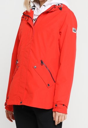 Persona che indossa una giacca arancione con cappuccio e tasche con zip e bottoni, sopra una felpa bianca con cappuccio e pantaloni neri.