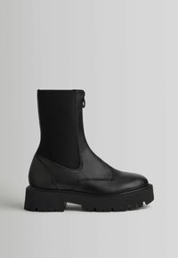 Bottine noire en cuir avec semelle crantée épaisse, panneaux latéraux élastiques, bout arrondi, et tirette de fermeture éclair sur fond gris uni.
