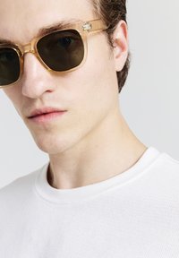 Giovane uomo che indossa occhiali da sole trasparenti marroni e una maglietta bianca a girocollo con texture, su uno sfondo chiaro.