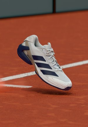 White athletic shoe with navy blue accents, textured upper, and distinctive three-stripe logo, floating above a red clay court surface.