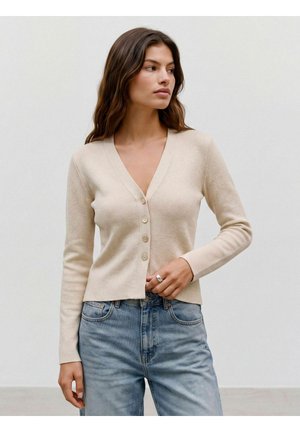 Femme aux longs cheveux bruns portant un cardigan beige boutonné et un jean bleu clair, regardant sur le côté devant un fond uni.