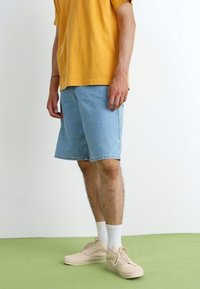 Denim shorts in light blue, featuring a relaxed fit and knee-length cut, paired with a mustard yellow t-shirt and beige sneakers.