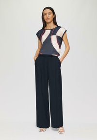 Marinebluse mit abstraktem Muster in Creme und Rot, kurze Ärmel; hochtaillierte, weit geschnittene schwarze Hose; weiße Riemchensandalen setzen einen Akzent bei der Fußbekleidung.