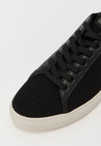 Svart sneakers med mesh-overdel og lærdetaljer, flate svarte snøring og hvit gummisåle. Minimalistisk design med rene linjer.