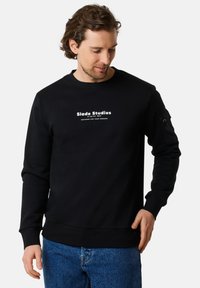 Svart sweatshirt med rundad halsringning, prydd med vit tryckt text på bröstet och en sidoficka med knappdetalj. Texturerad tyg.