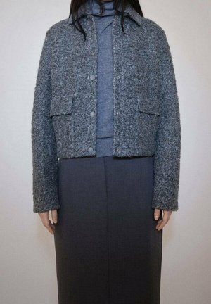 Femme portant une veste grise texturée boutonnée sur un pull à col roulé bleu et une jupe gris foncé sur un fond uni.