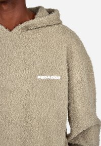 Mikina s kapucí ve světle béžové barvě, vyrobená z texturované tkaniny. Obsahuje viditelné bílé logo "PEGADOR" na hrudi.
