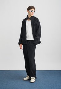 Conjunto de casaco e calças de flanela preta com colarinho com fecho éclair, combinado com camisa branca e óculos de sol prateados. Os sapatos têm um design preto e branco.