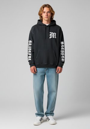 Felpa nera con scritte bianche sulle maniche e sul petto, abbinata a jeans in denim azzurri chiari e sneakers bianche con strisce nere.