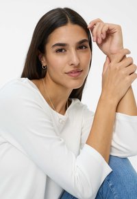 Weißes langärmliges Oberteil mit runder Ausschnitt, getragen zu blauer Jeans. Das Model hat glattes, dunkles Haar und kleine Creolen, die Arme liegen in der Nähe des Gesichts.