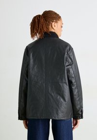 Giacca in pelle nera con una vestibilità rilassata, colletto alto e polsini con bottone. Presenta una texture liscia e un design minimalista.