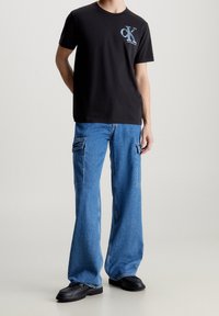 Černé bavlněné tričko s modrým grafickým logem, spárované s širokými modrými denimovými cargo kalhotami s bočními kapsami, doplněné černými botami.