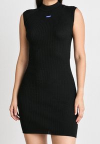 Robe noire côtelée avec un col montant et un design sans manches. Présente une petite étiquette logo bleue sur la poitrine. Silhouette ajustée.