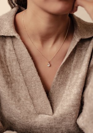 Gold-tone necklace featuring a pear-shaped clear crystal pendant, worn over a beige, textured knit sweater with a V-neck design.