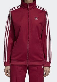 Rote Trainingsjacke mit weißen Streifen an den Ärmeln, hohem Kragen und Reißverschluss vorne; aus strukturiertem Stoff gefertigt; mit kleinem weißen Logo auf der Brust.