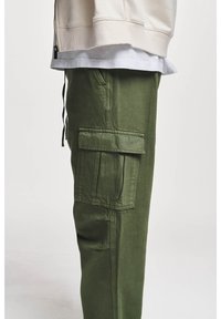Pantaloni cargo in verde oliva, con tasche laterali, vestibilità rilassata e tessuto strutturato. Indossati con un felpa color crema sopra una camicia bianca.
