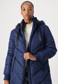 Manteau matelassé marine avec un motif en chevrons, fermeture éclair sur le devant et capuche ajustable. Doté de quincaillerie noire et de poignets côtelés pour plus de chaleur.