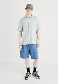 Lys grå kortermet skjorte med knapper, blå denimshorts og flerfargede joggesko. Kappen er olivengrønn med et logoakent.
