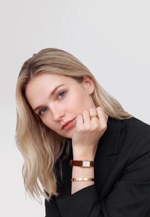 Vrouw met blond haar, gekleed in een zwart colbert, een horloge met een bruine leren band, een gouden armband en een gouden ring, die haar kin op haar hand laat rusten.