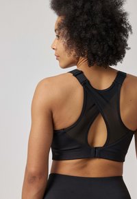 Reggiseno sportivo nero con dettagli in rete, design a incrocio sulla schiena, spalline regolabili e chiusura con gancio sul retro. Texture liscia e vestibilità supportante.