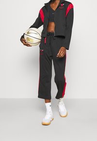 Sort atletisk tøj med røde accenter, der inkluderer en lynlåsjakke og cropped bukser. Modellen holder en hvid basketball med et Nike-logo.