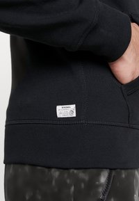 Felpa nera con tessuto testurizzato, cuciture visibili e una piccola etichetta bianca del marchio vicino all'orlo. Presenta polsini e orlo a costine.