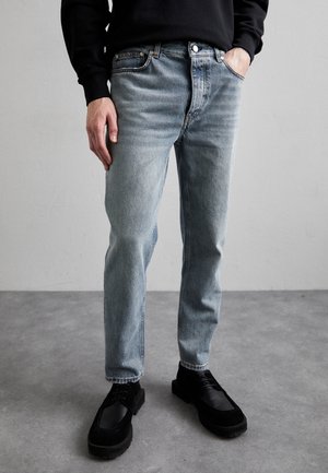 Mężczyzna ubrany w jasnoniebieskie dopasowane dżinsy, czarne sznurowane buty oraz czarną koszulę z długim rękawem, z jedną ręką w kieszeni.