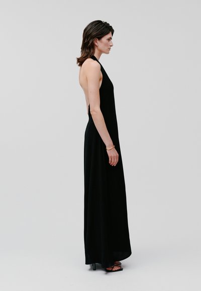 Black halter-neck maxi dress in a fluid fabric, featuring an open back and a sleek silhouette. Paired with black heeled sandals.