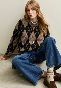 Jeune femme aux longs cheveux blonds portant un pull en argyle marron et noir, un jean large bleu, des chaussettes beiges et des mocassins marron, assise sur le sol.