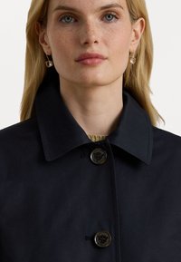 Abrigo azul marino con cuello clásico, dos botones oscuros y una textura suave. Pendientes dorados con acentos transparentes completan el look.