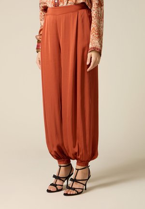 Donna che indossa pantaloni larghi color ruggine con polsini arricciati e sandali neri con tacco a cinturini, in piedi su uno sfondo neutro.