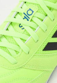 La zapatilla deportiva en verde neón presenta materiales de malla y ante, cordones planos, un logo negro y detalles texturizados en la parte superior y en la suela.