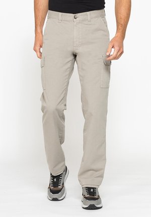 Uomo che indossa pantaloni cargo beige chiaro e sneakers nere e grigie che cammina su sfondo bianco.