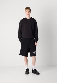 Black crewneck sweatshirt and black basketball-style shorts with a printed logo in white. Model wears black sneakers and white socks.