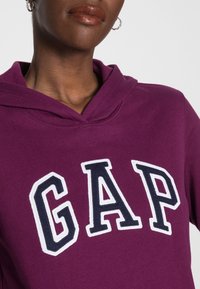 Fialová mikina s kapucí a bílými a námořnicky modrými písmeny tvořícími nápis "GAP". Měkká látka s texturovaným povrchem, vybavená klokáním kapsou a stahovací šňůrkou.