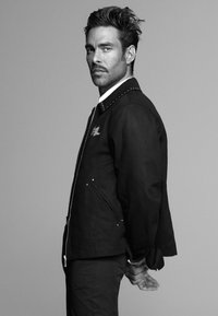 Melna jaka ar sudraba elementi un studētu apkakli, ar rāvējslēdzēja aizdari un sānu kabatām, uzvilkta virs baltas krekla.