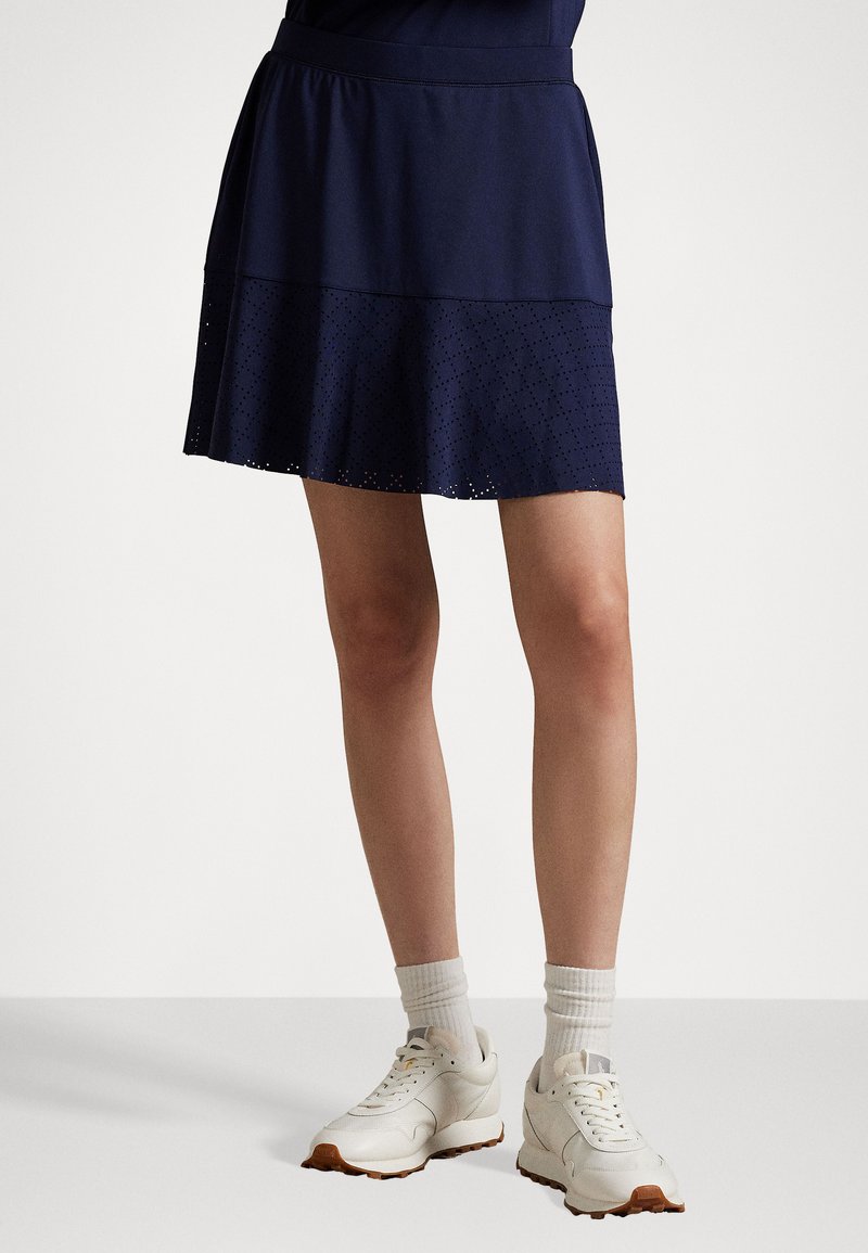 Marineblå skorts med lagdelt design; øverste halvdel er ensfarget, mens den nederste halvdel har et perforert mønster. Kombinert med lyse joggesko og hvite sokker.