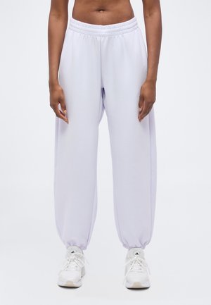 Personne portant un pantalon de survêtement violet clair à taille et chevilles élastiques avec des baskets blanches Adidas sur un fond blanc uni.