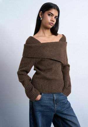 Ung kvinde med langt mørkt hår iført brun off-shoulder ribbet sweater og blå jeans, hænderne i lommerne, neutral baggrund.